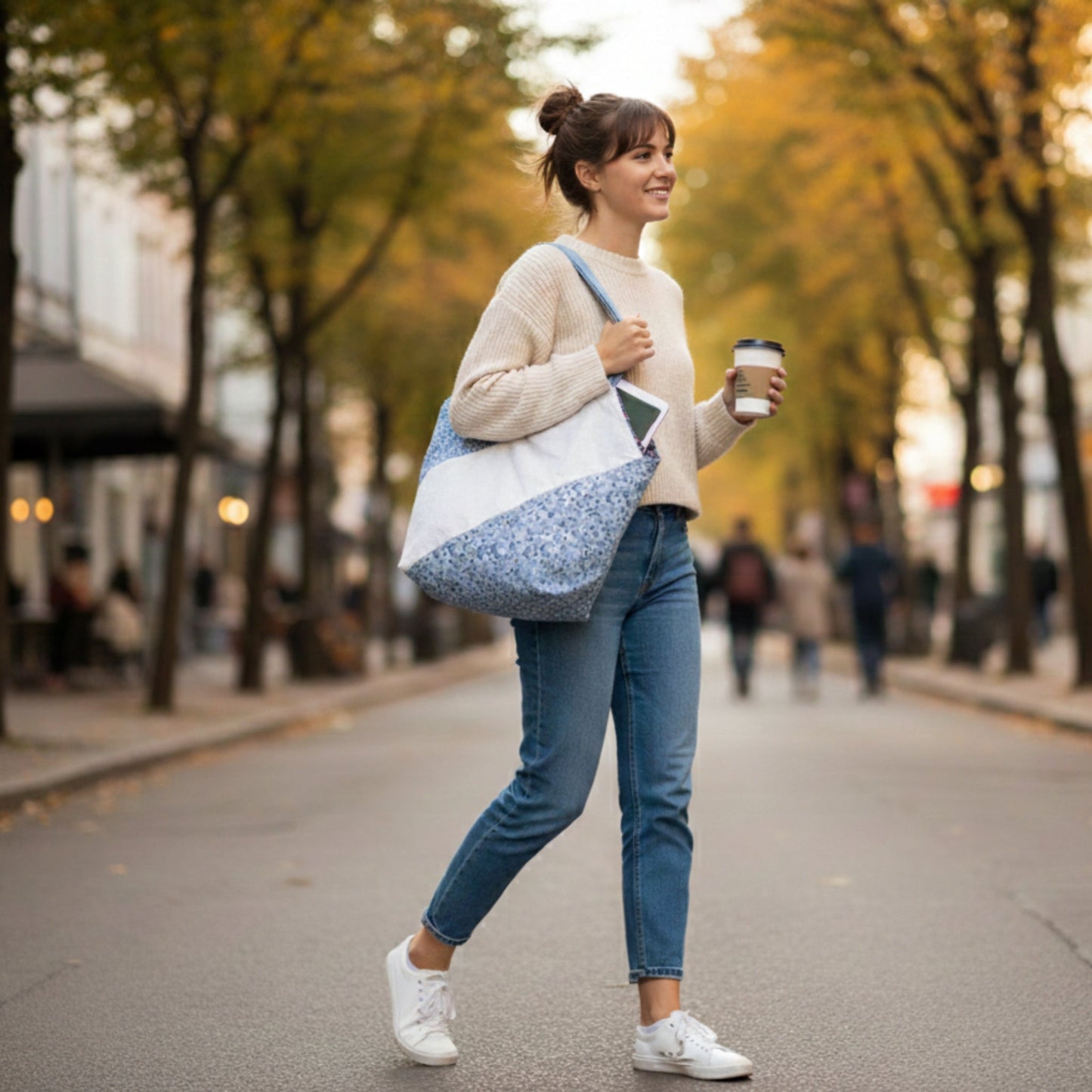 Full-body shot of a young woman walking down a city street with tree-lined sidewalks, carrying the Blue Floral Tote Bag over her shoulder. The bag features blue floral print and a white diagonal stripe. She is wearing jeans, a beige sweater, and white sneakers, highlighting the tote's light, casual style for daily errands