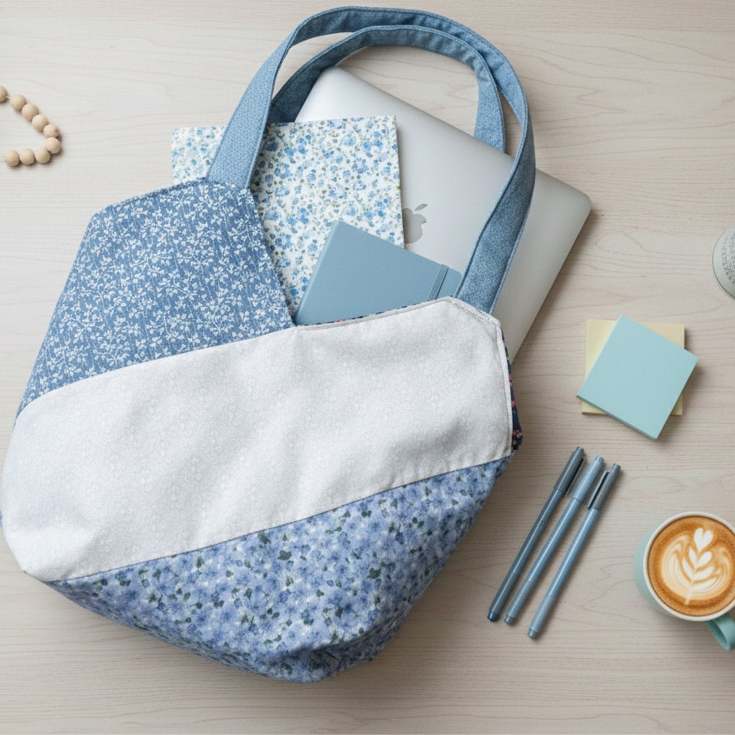 Lifestyle flat lay of the Blue Floral Tote Bag on a light wood desk, filled with a laptop, notebooks, and a planner. The exterior features a blue floral print and a white diagonal stripe. Pens, sticky notes, and a cup of latte sit nearby, illustrating the tote's generous capacity for work or school essentials