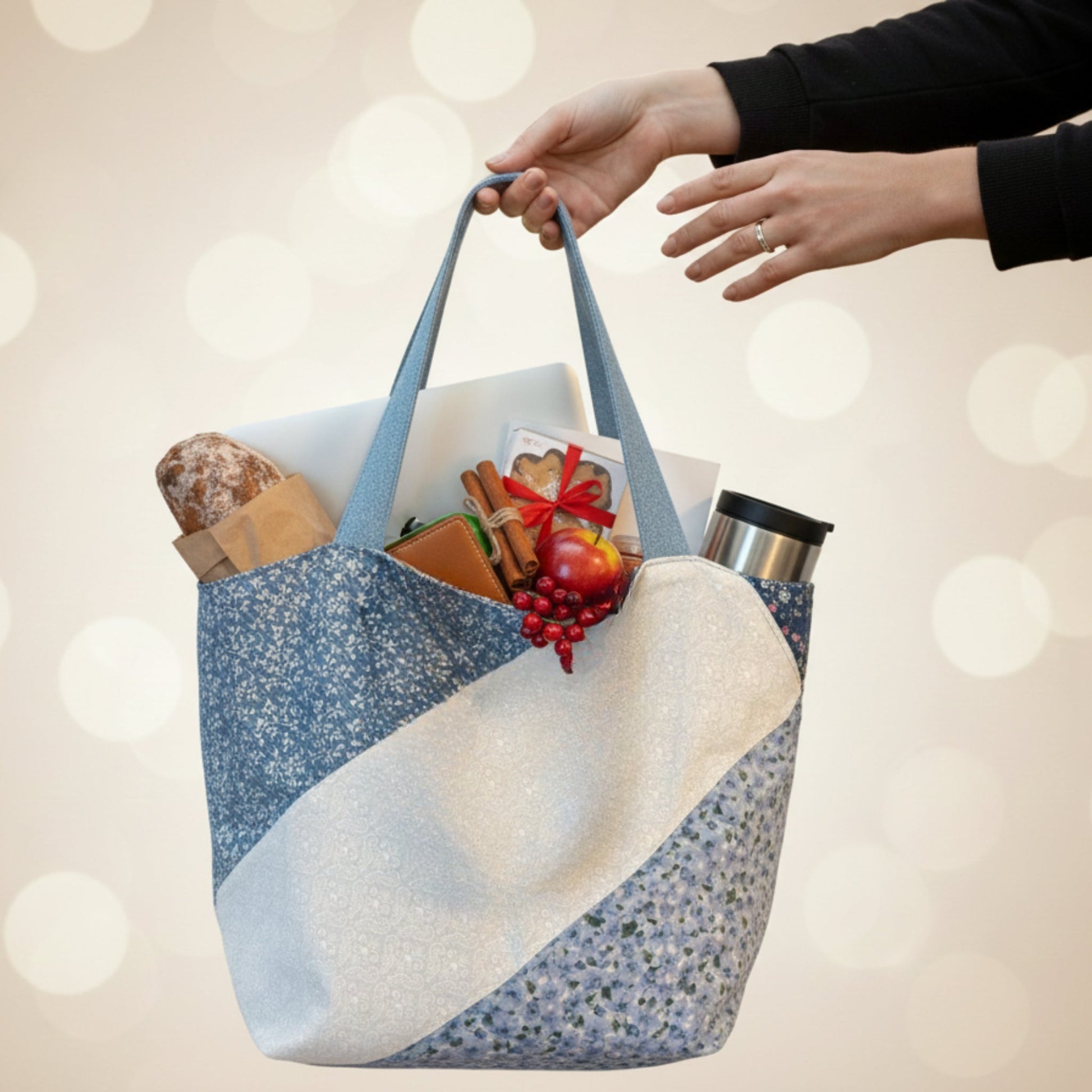 Studio shot of two hands holding up the Blue Floral Tote Bag against a gold bokeh background. The bag is filled with a loaf of bread, a laptop, and holiday-themed items like an apple and cinnamon sticks, showing its versatility for shopping, work, or gift-giving