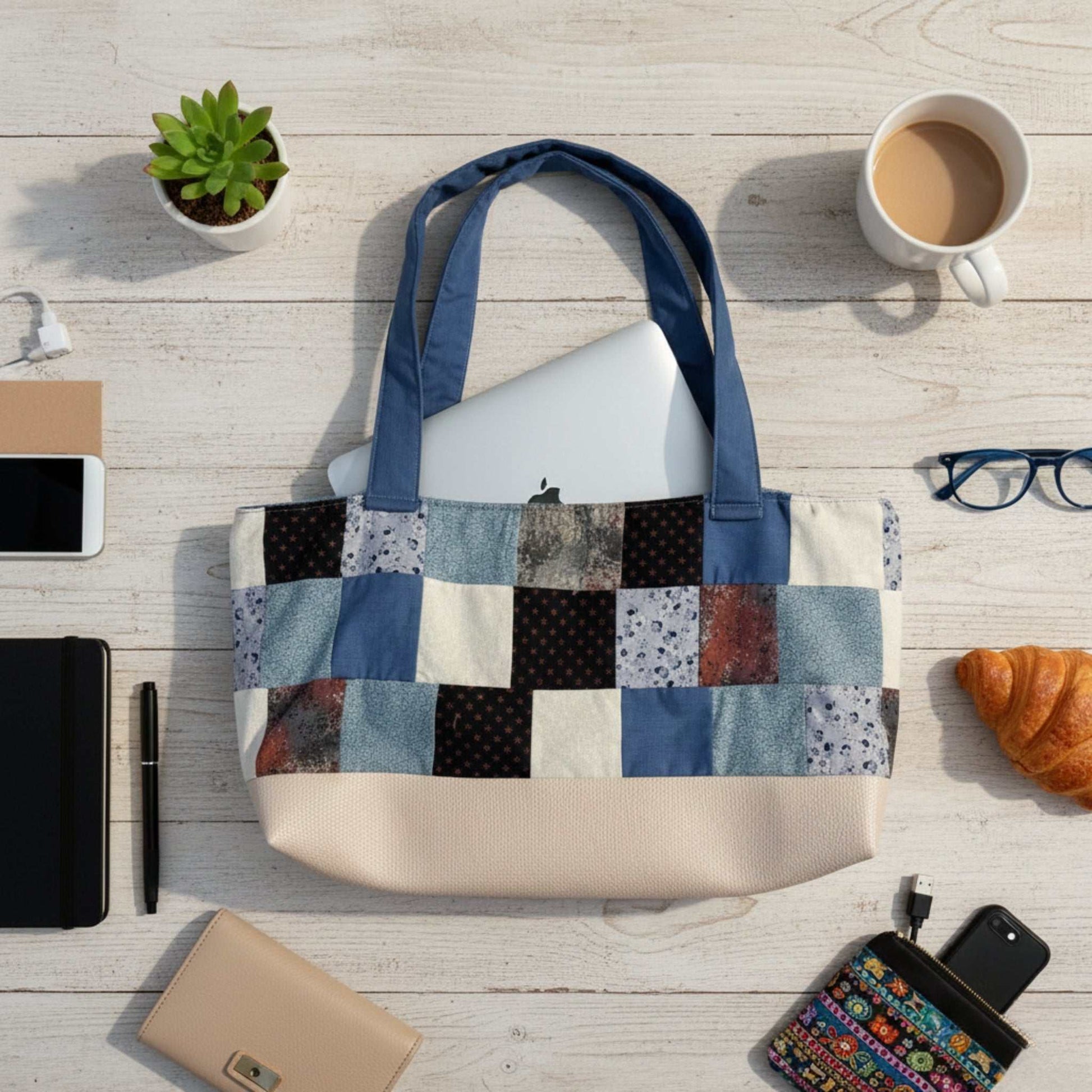 Flat lay shot of the Handmade Patchwork Tote Bag on a white wooden desk, filled with a silver laptop. The tote has a cream faux-leather base and patchwork squares in various blue, cream, and dark red fabrics. It is surrounded by a croissant, coffee, glasses, and a wallet, demonstrating the bag's capacity for daily work essentials.
