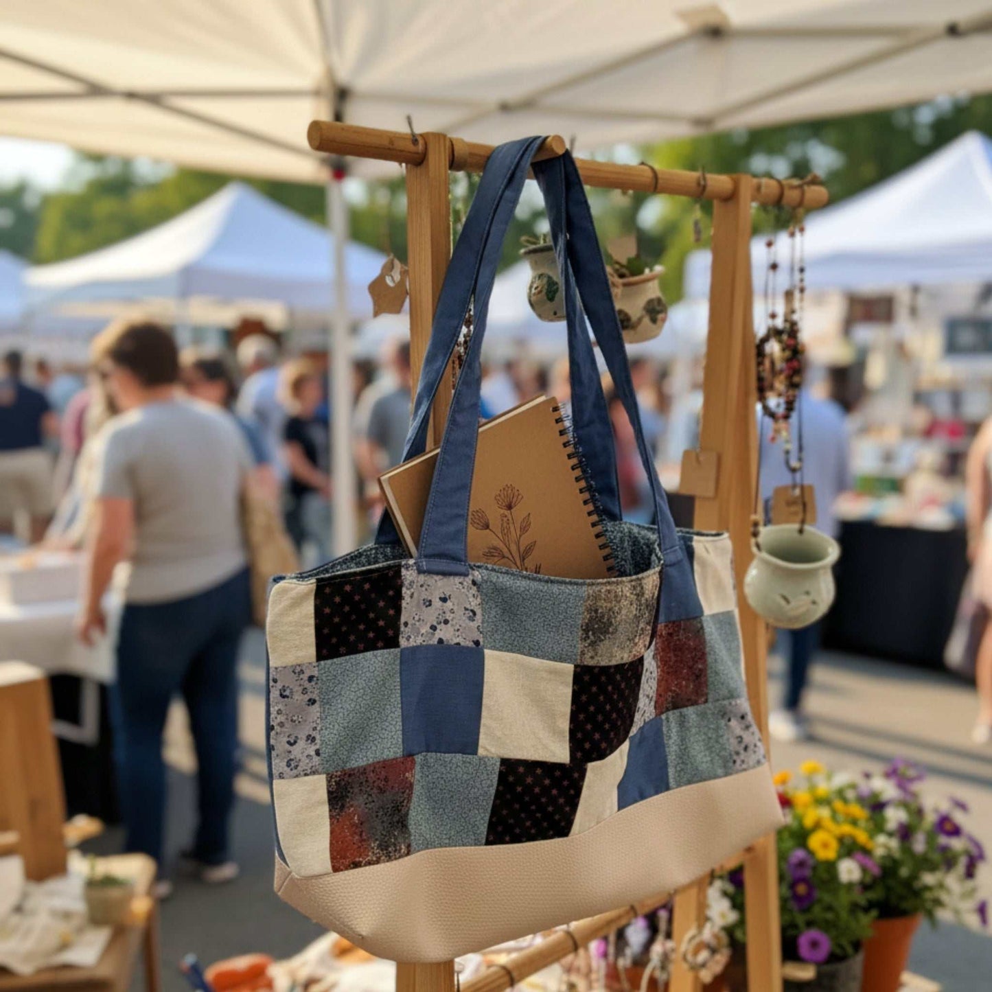 Handmade Patchwork Tote Bag in shades of blue, cream, and rustic red hanging outdoors at a market stall. The bag has a cream faux-leather bottom and blue denim straps. It is stuffed with a notebook, showing its use for market shopping or crafts.