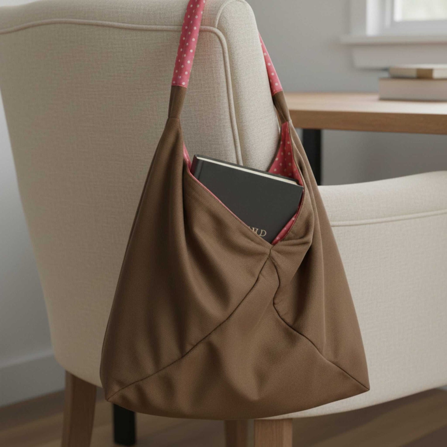 Close-up of the Reversible Slouch Hobo tote Bag  in its solid taupe exterior, hanging on a chair. A dark book is tucked into the large exterior pocket, revealing the pink polka dot lining on the strap, which demonstrates its dual-color design