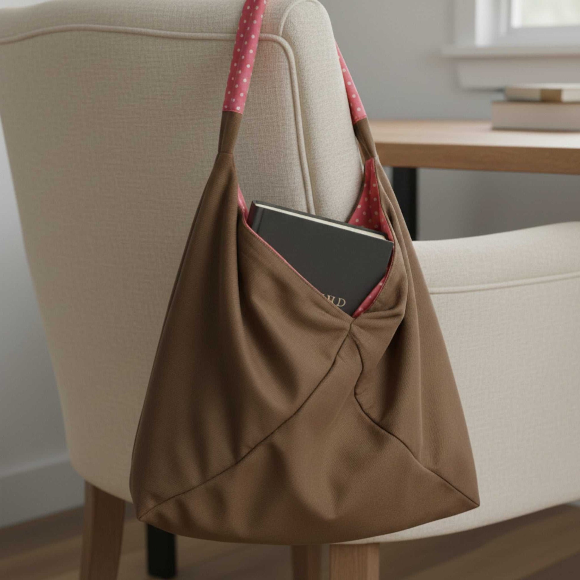 Close-up of the Reversible Slouch Hobo tote Bag  in its solid taupe exterior, hanging on a chair. A dark book is tucked into the large exterior pocket, revealing the pink polka dot lining on the strap, which demonstrates its dual-color design