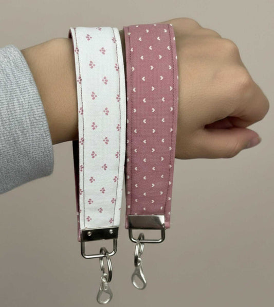 Close-up shot of a hand modeling two patterned fabric Keychain wristlets: one in white with pink accents, and one in dusty rose pink with white hearts. Both wristlets feature a metal clip for attaching keys or a wallet.