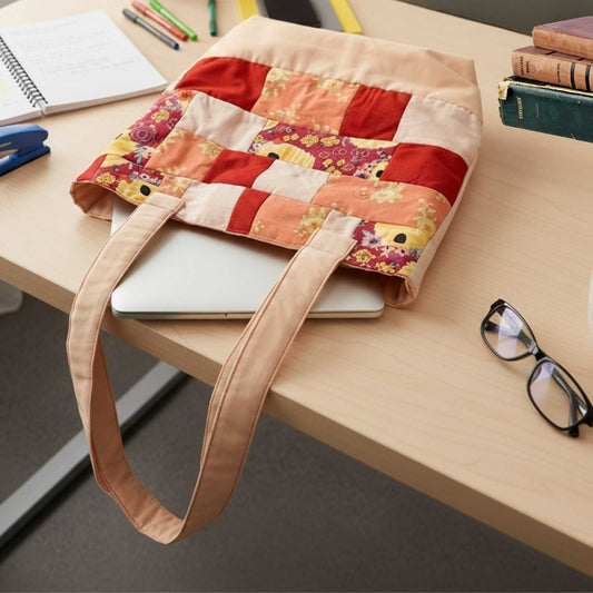 Flat lay of the Red and pink Handmade Patchwork Tote Bag on a wooden desk, placed over a silver laptop. The tote is surrounded by books, glasses, and a stapler, demonstrating its capacity for school or office supplies.