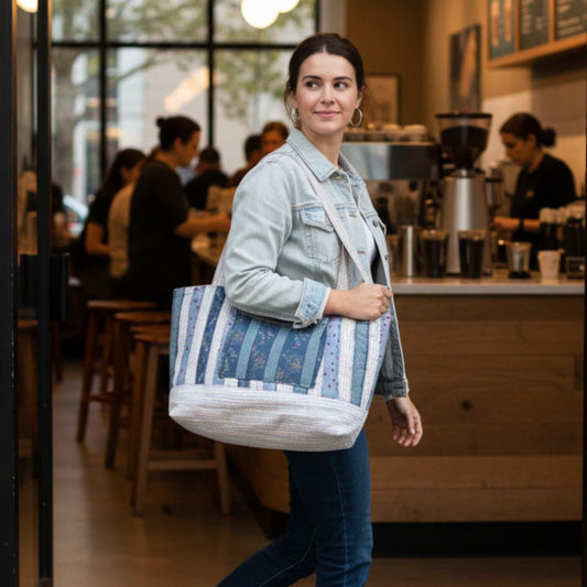 A woman wearing a light denim jacket and jeans carries a large, handmade quilted tote bag with blue, white, and striped fabric patterns over her shoulder as she walks through the entrance of a busy coffee shop.